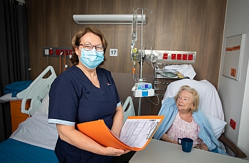 A nurse in mask standing with an file next to a old lady in an hospital bed